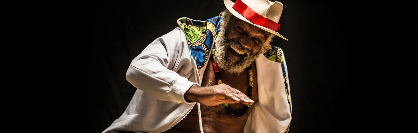 Mestre Lumumba sorri com expressão de alegria, vestindo chapéu branco com uma faixa vermelha.