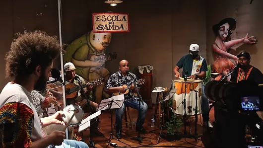 Grupo de músicos em roda de samba, com diversos instrumentos de percussão e cordas.