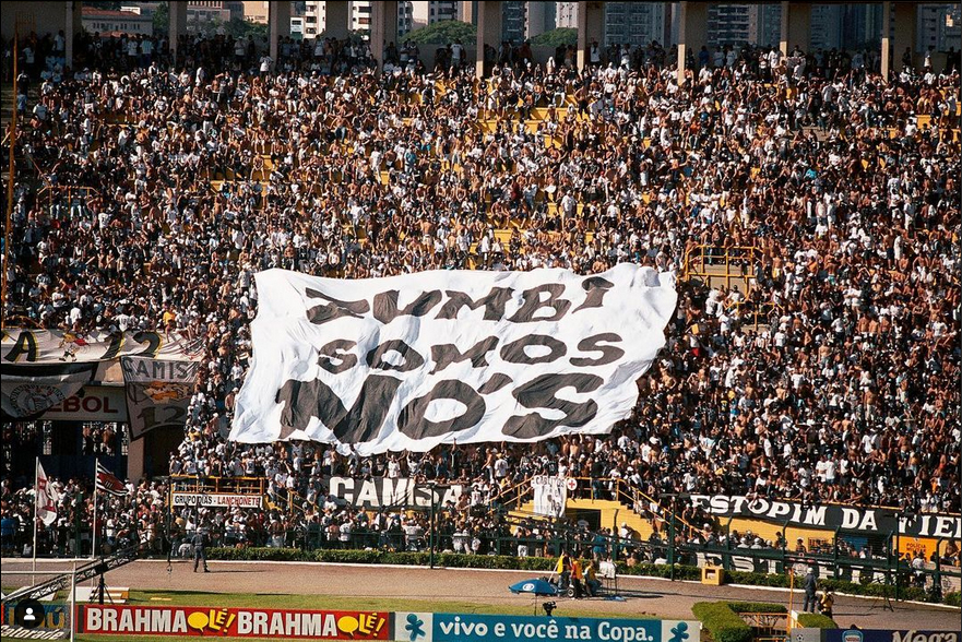 Grande multidão segurando uma faixa com a frase "ONDE ESTÃO OS NEGROS?" em um estádio de futebol, com uma partida ocorrendo. A faixa é branca com texto em letras pretas.