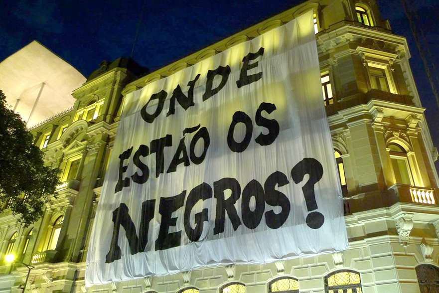 Grande faixa com a pergunta "ONDE ESTÃO OS NEGROS?" pendurada em um prédio art déco em São Paulo durante a noite.