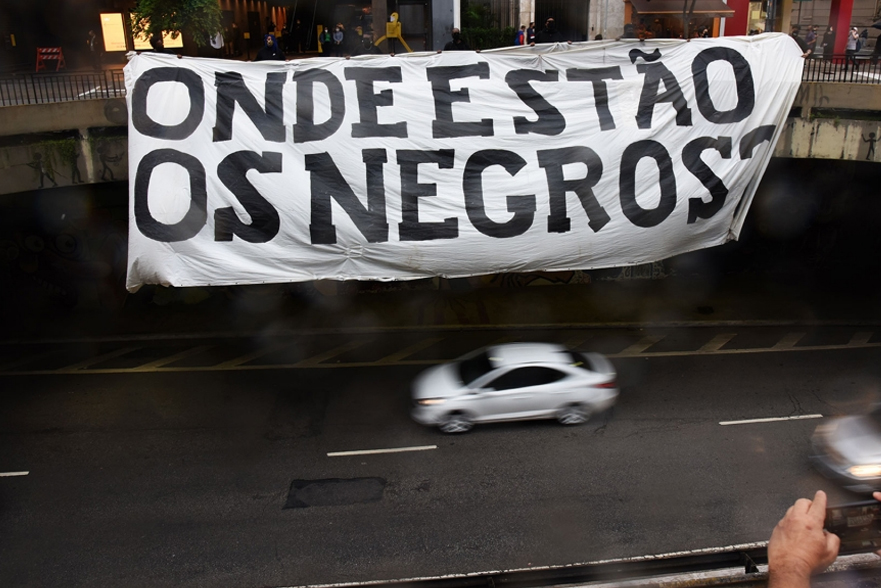 Faixa branca com a frase "ONDE ESTÃO OS NEGROS?" escrita em letras pretas, sendo segurada por pessoas em um viaduto acima da rua.