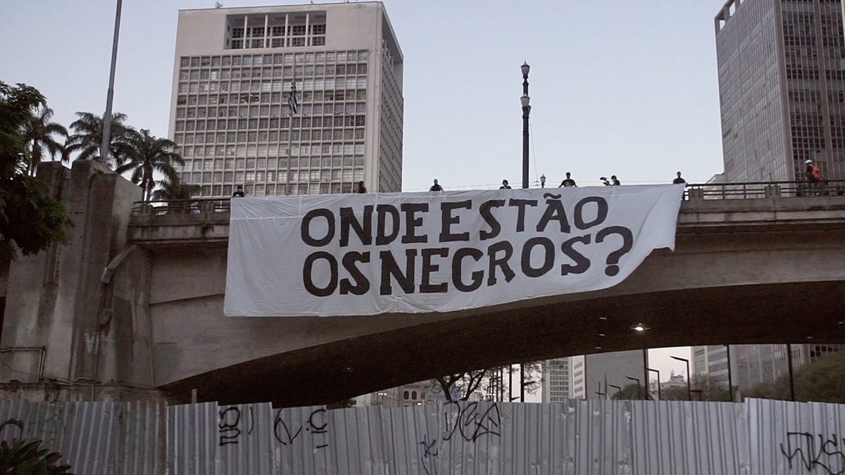 Faixa branca com a pergunta "ONDE ESTÃO OS NEGROS?" pendurada em um viaduto durante o dia.