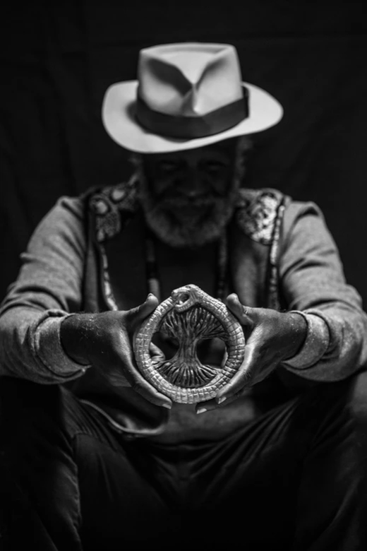 Fotografia em preto e branco do Mestre Lumumba usando chapéu, segurando as mãos à frente, destacando um Ouroboros antigo.