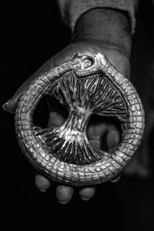 Fotografia em preto e branco de um Ouroboros antigo em forma de uma cobra comendo a própria cauda com uma árvore no centro, segurado pelo Mestre Lumumba. O metal possui desenhos intrincados que lembram raízes ou galhos.