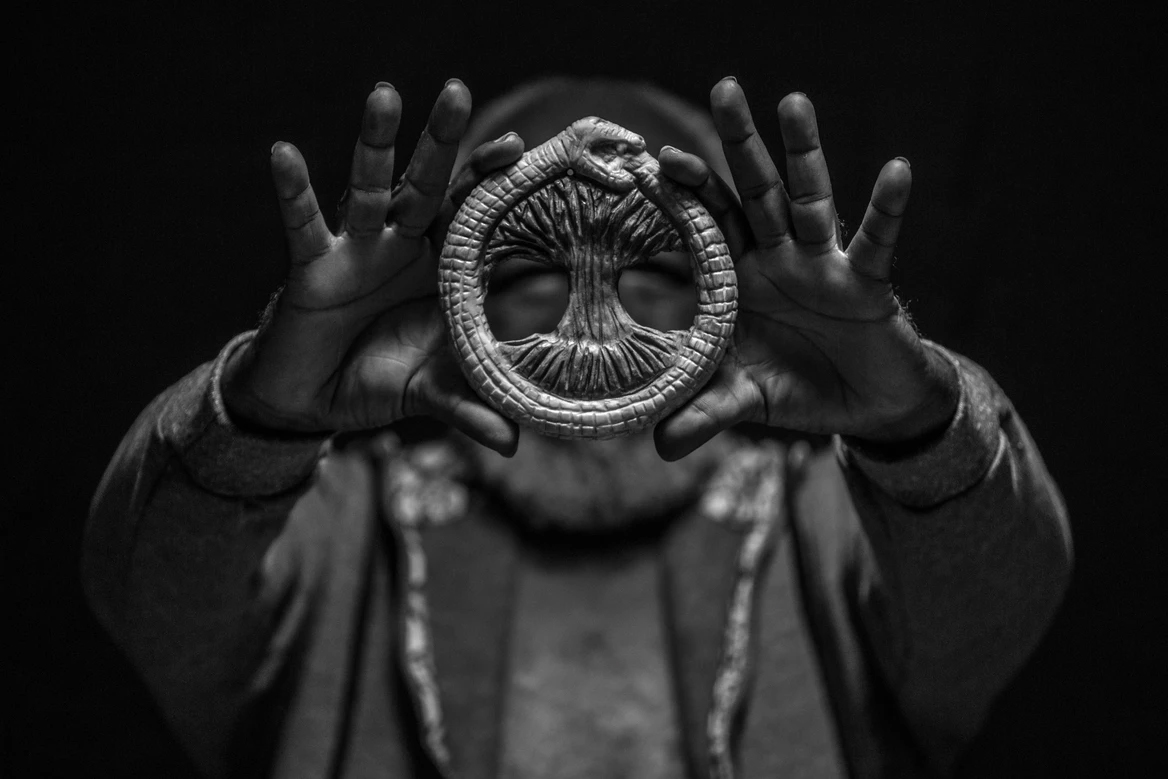 Fotografia em preto e branco de um homem com as mãos levantadas, segurando um Ouroboros antigo em forma de uma cobra comendo a própria cauda com uma árvore no centro, contra um fundo escuro e texturizado.