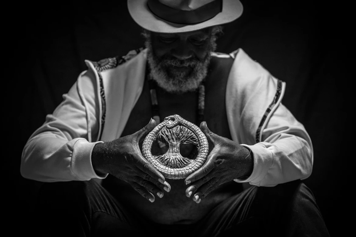 Fotografia em preto e branco do Mestre Lumumba usando chapéu branco, segurando um Ouroboros, de uma cobra comendo a própria cauda com uma árvore no centro.